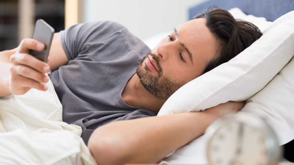 A close up shot of a man lying in bed immediately reaching for his smartphone upon waking up This image illustrates the morning phone reach a dangerous habit that triggers dopamine spikes and contributes to neural burnout by disrupting the brain's natural waking process