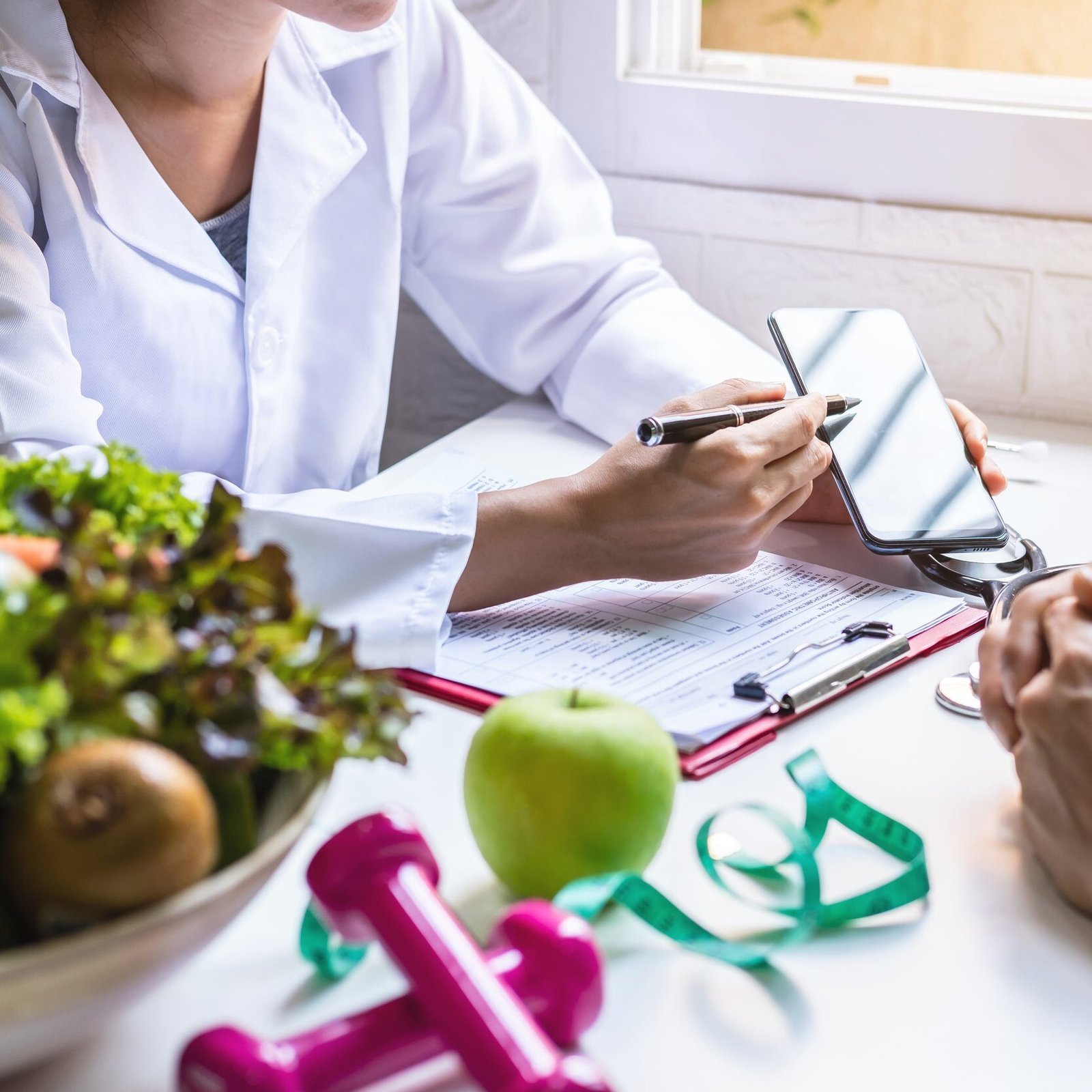 A close up view of a nutritionist in a white coat providing a personalized metabolic wellness consultation using a smartphone to explain a plan alongside fresh green vegetables a green apple and pink dumbbells for physical activity resistance
