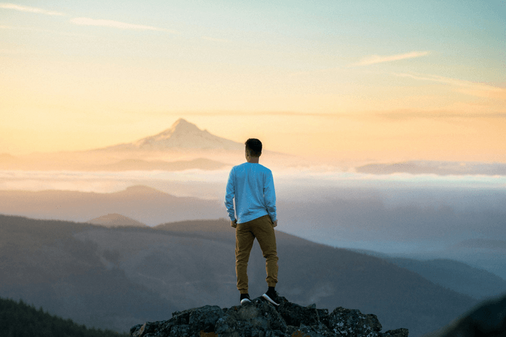 A man standing on a high mountain peak at sunset representing the journey to overcome neural burnout and achieving a state of peak focus and mental clarity through deep rest and nature