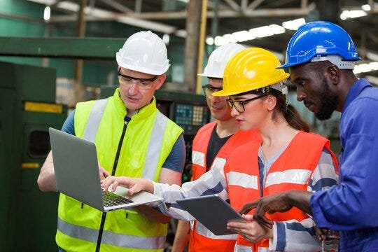 Four industrial workers wearing hard hats, safety glasses, and high-visibility vests collaborate in a factory, reviewing information on a laptop and tablet during a production floor meeting.