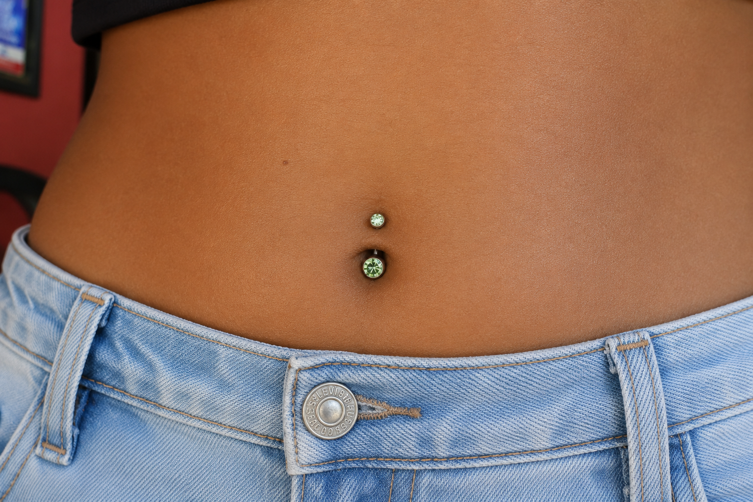 A close-up of a lower navel piercing with green gemstone curved barbell jewelry, used to compare jewelry styles when researching what is a fourchette piercing.