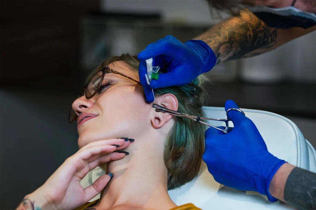 A professional piercer wearing blue medical gloves uses a sterile needle and clamps on a client, illustrating the safety standards to look for when researching what is a fourchette piercing.