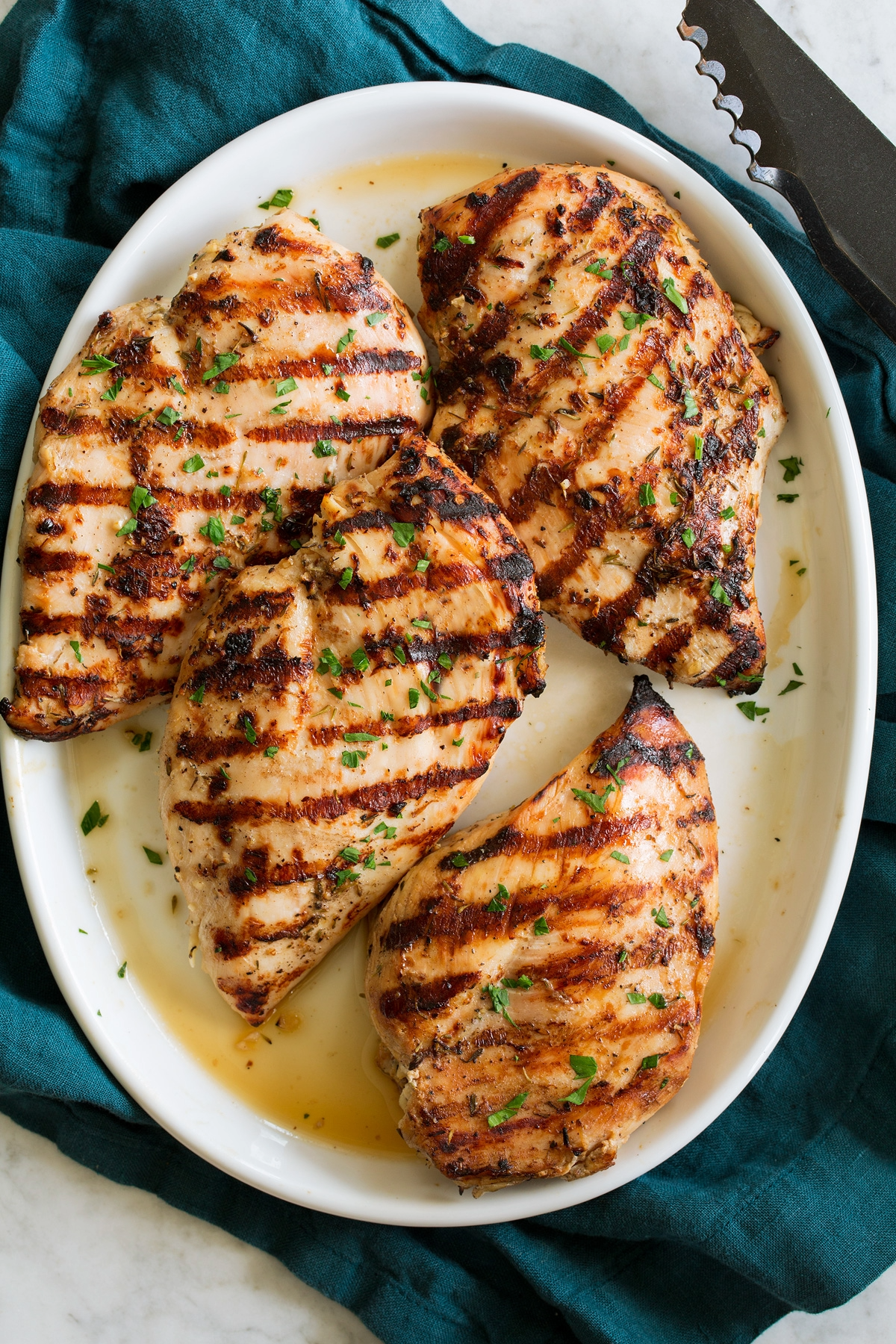 Four herb marinated grilled chicken breasts with visible grill marks and fresh parsley garnish on a white oval plate